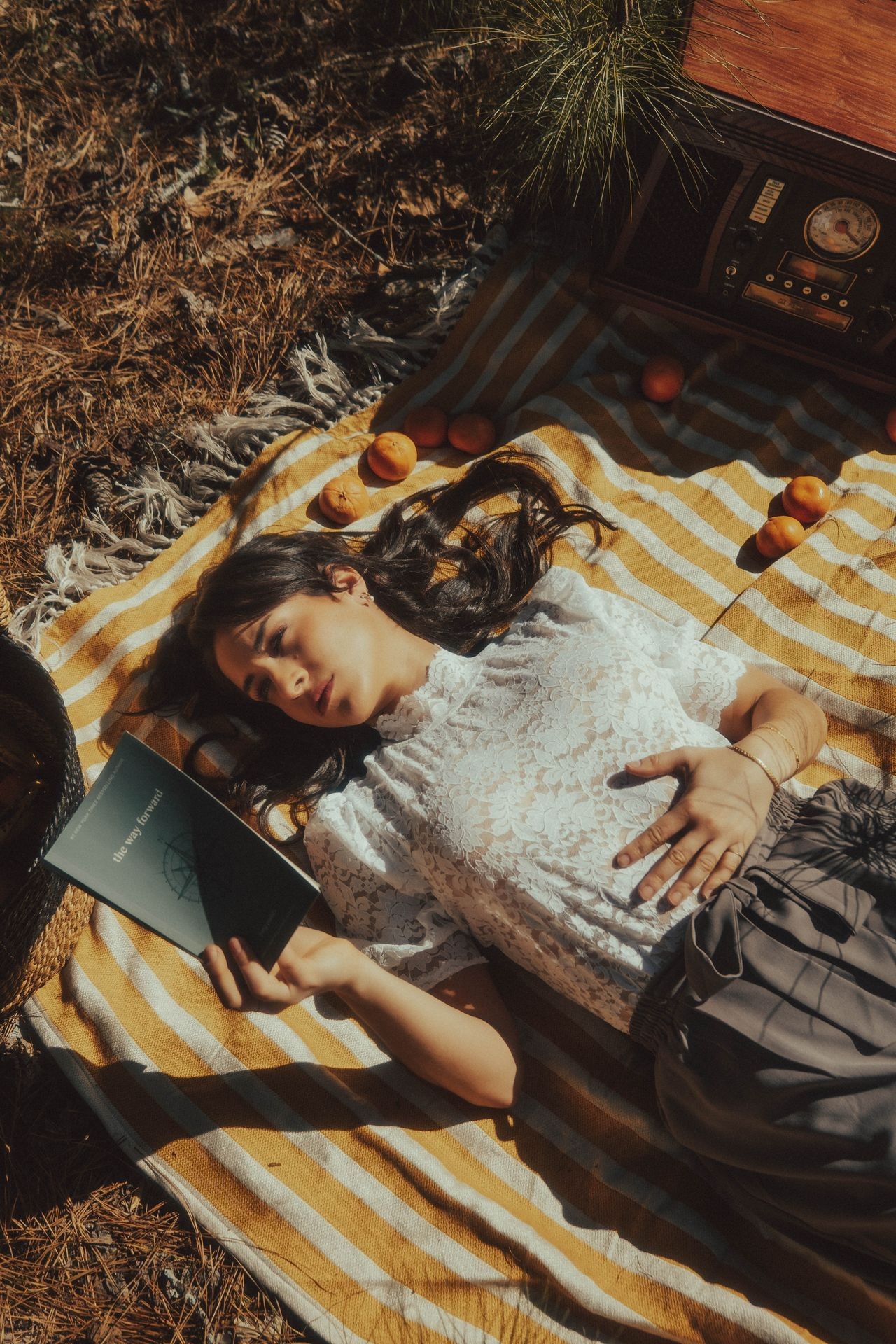 Woman lying on a striped blanket outdoors, holding a book, with oranges scattered around her.