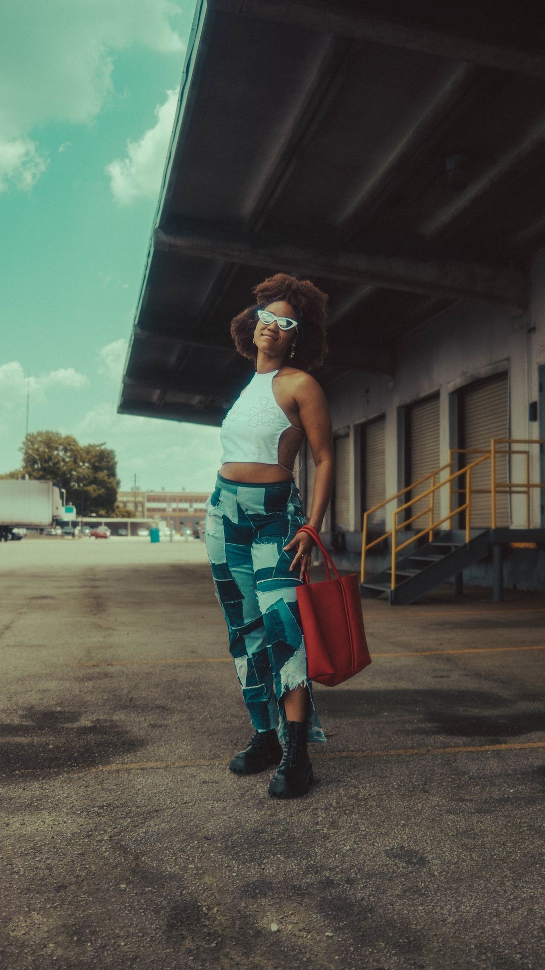 Fashionable person with sunglasses and patchwork jeans posing outdoors holding a red bag.