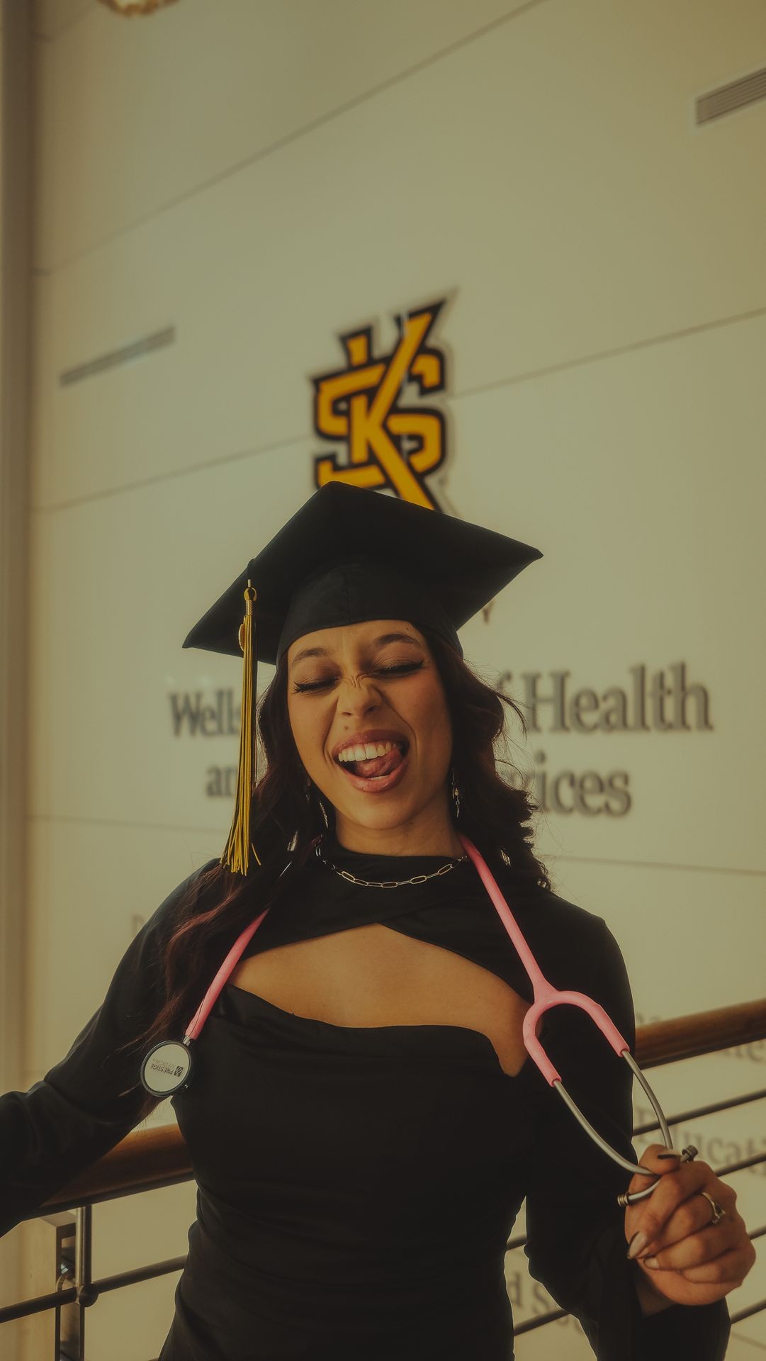 Graduate in cap with stethoscope posing playfully indoors, wearing a gown and smiling.