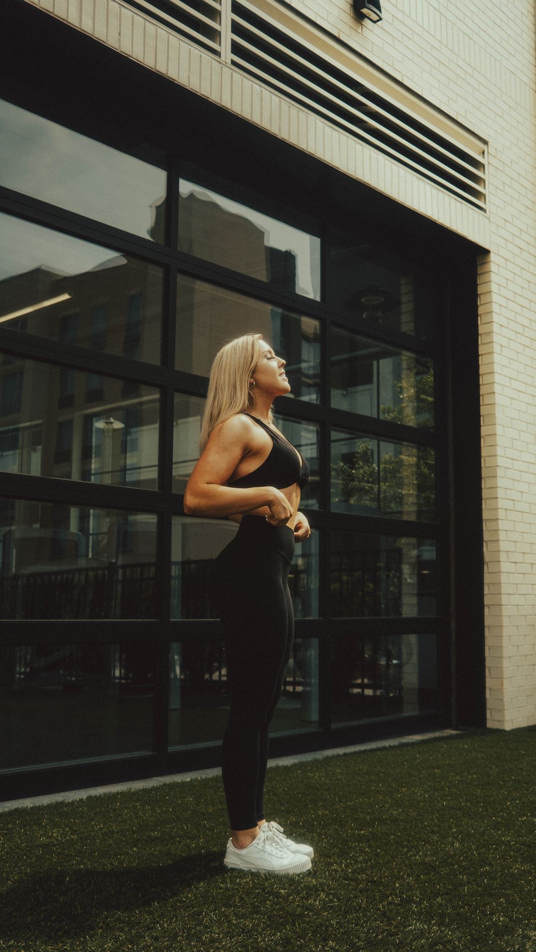 Woman in workout attire stands confidently on artificial grass in front of a modern glass building.